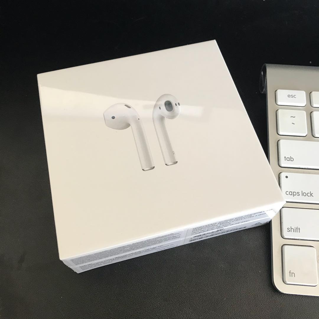 White wireless earbuds box on a black desk beside part of a white keyboard.