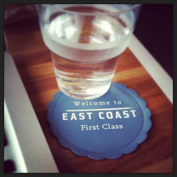 Plastic cup of water on a blue coaster reading “Welcome to East Coast First Class” on a wood table.