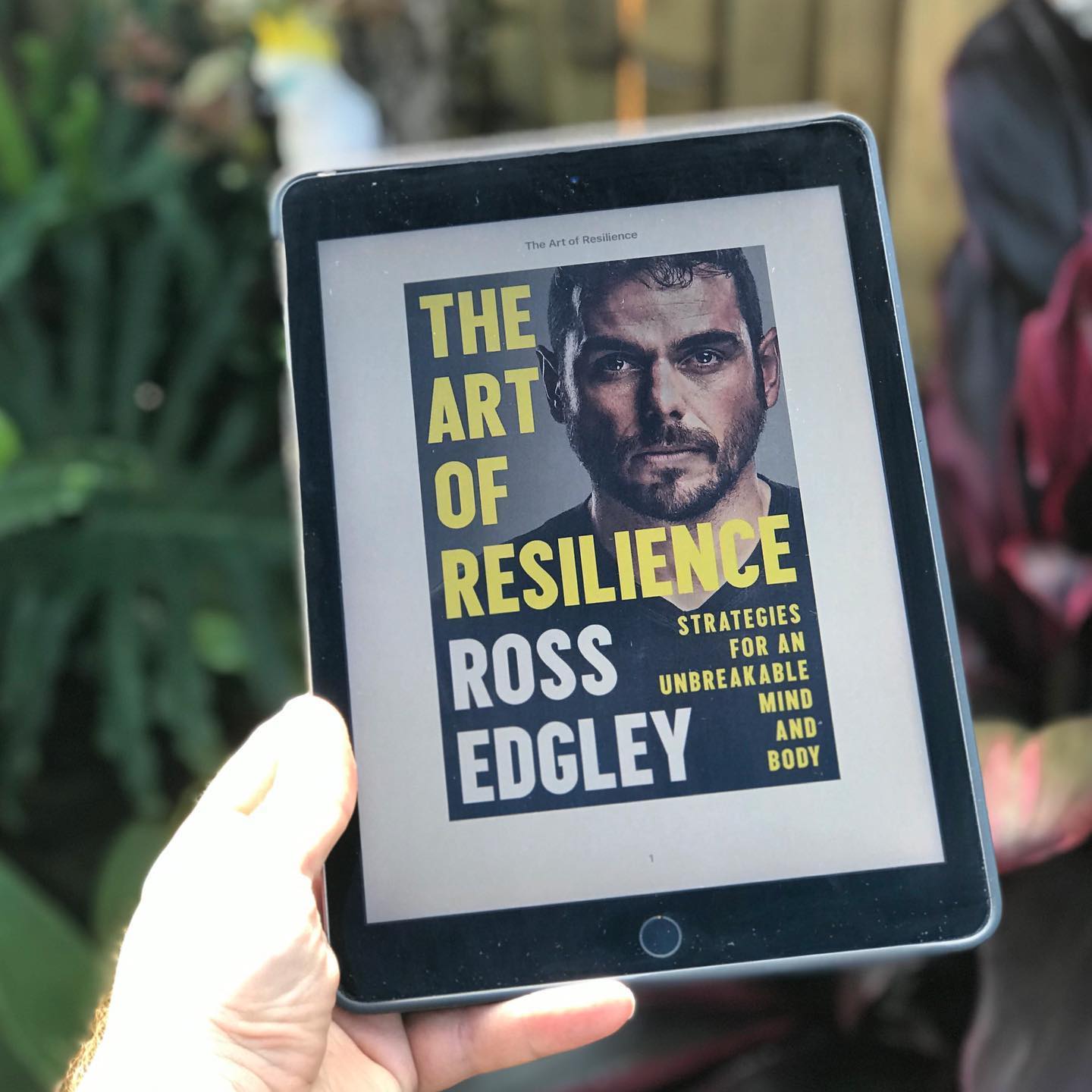 Hand holding a tablet displaying the book cover "The Art of Resilience" by Ross Edgley against a blurred green background.
