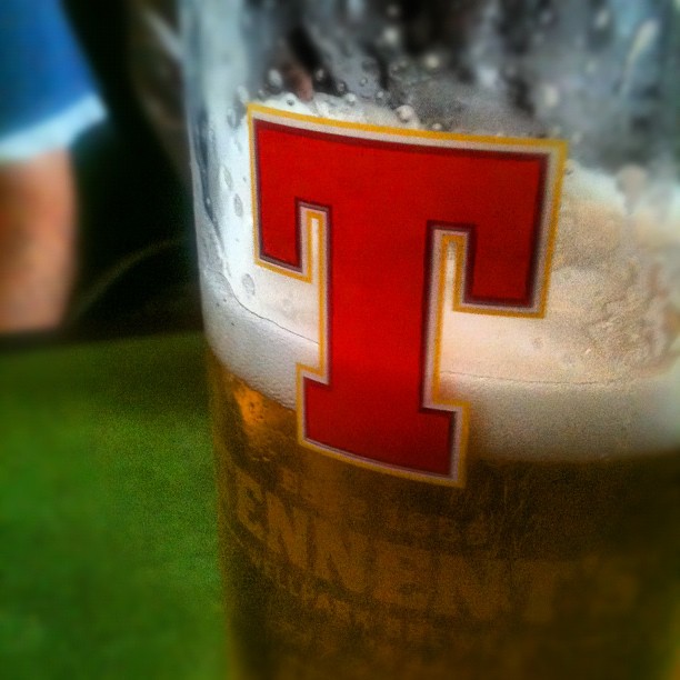Close-up of a partially full beer glass with foam and a large red letter T on the side.