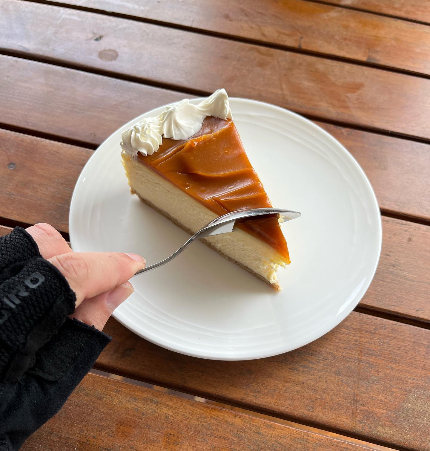 Slice of cheesecake with caramel topping and whipped cream on a white plate, with a hand holding a fork over it on a wooden table.