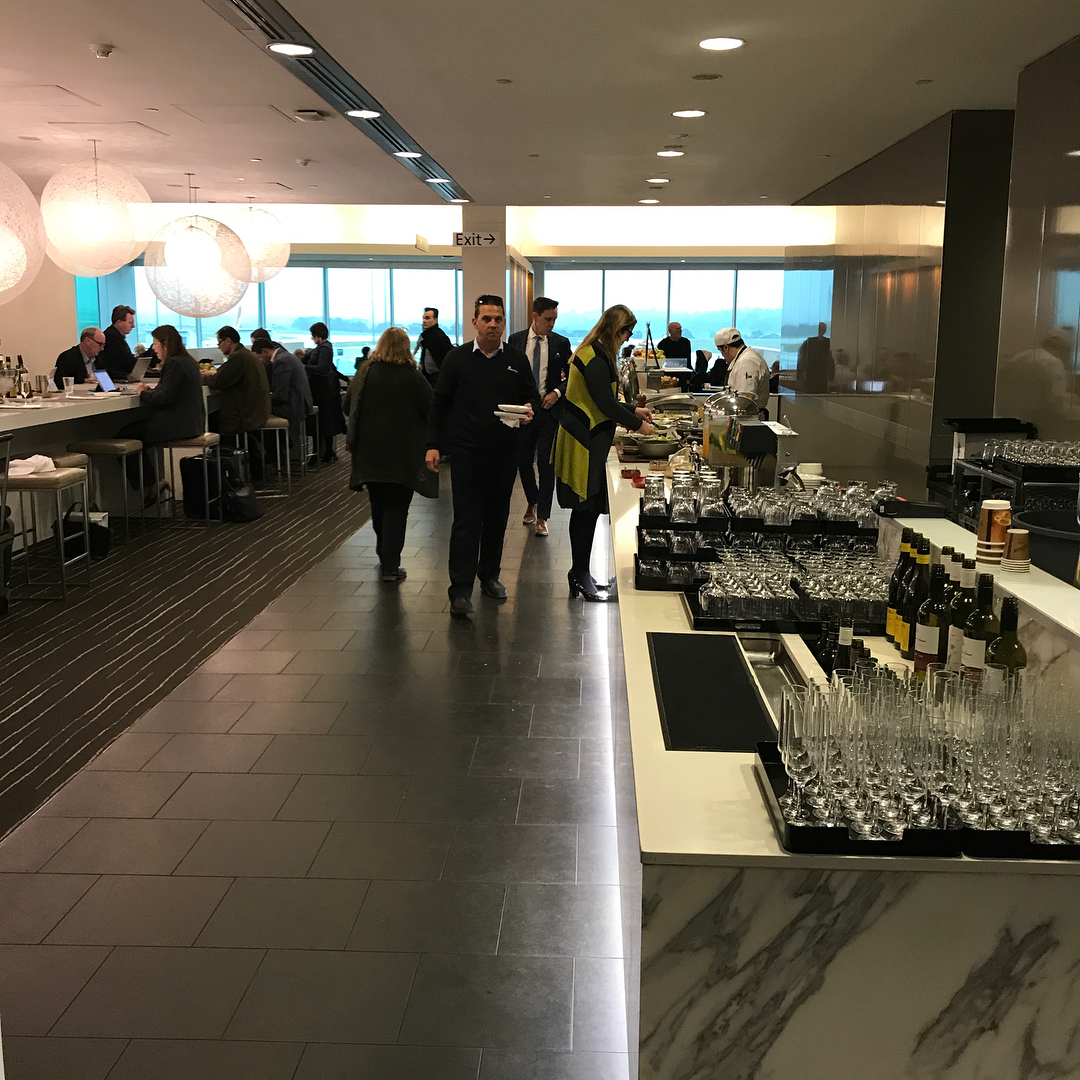 Airport lounge interior with seated guests, a buffet line, and a counter filled with glasses and wine bottles.