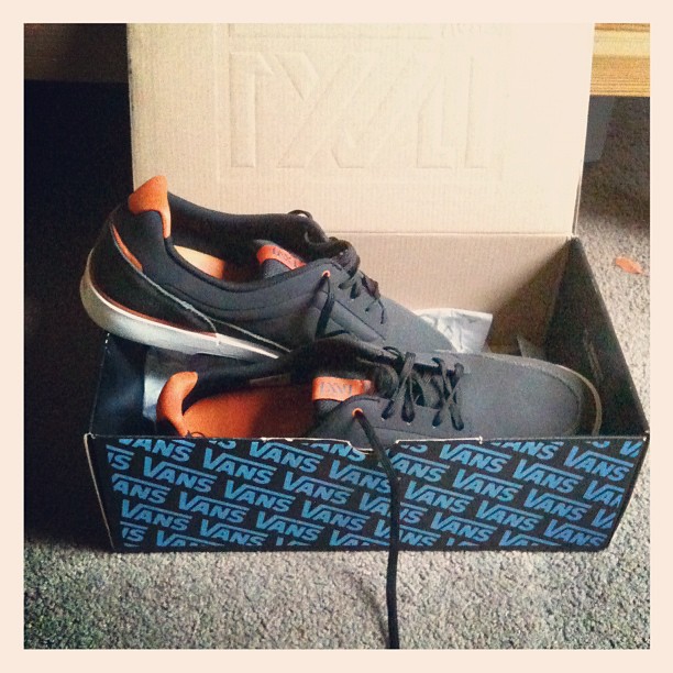 Black and orange Vans sneakers in an open shoebox on a carpeted floor.