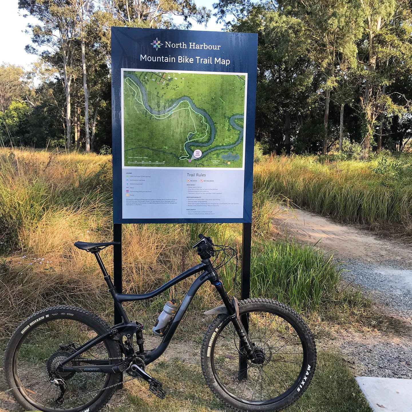 Mountain bike parked in front of a North Harbour mountain bike trail map beside a dirt path and tall grass.