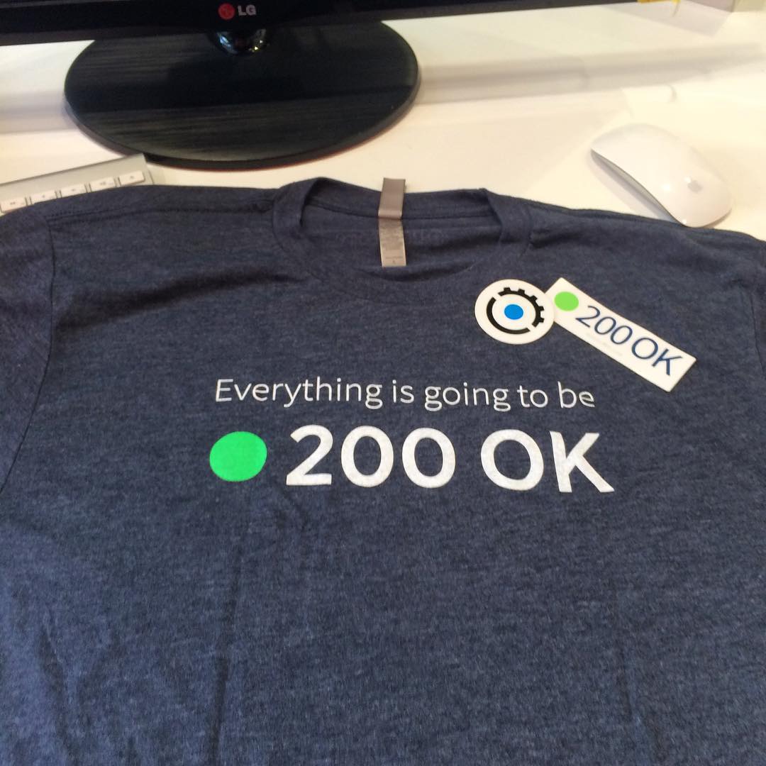 Blue T-shirt on a desk reading “Everything is going to be 200 OK,” with matching stickers near the collar.