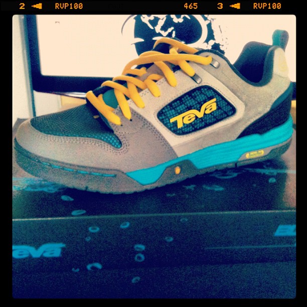 Gray and teal Teva sneaker with yellow laces displayed on a black shoebox in front of a bright screen.