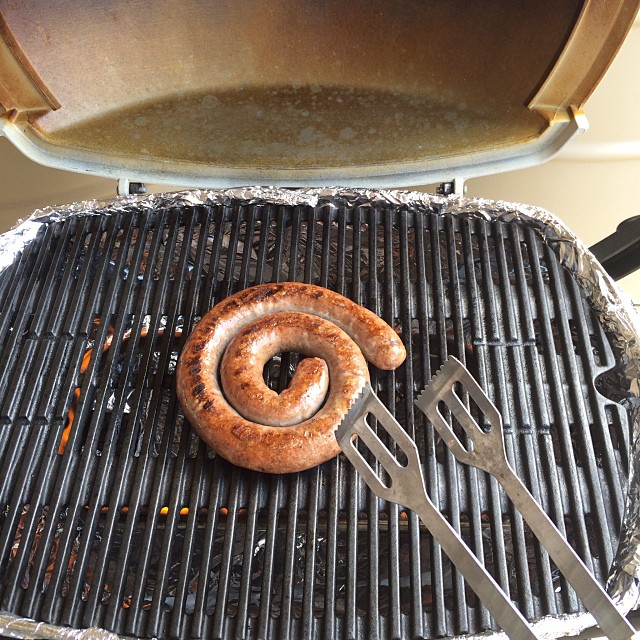 Coiled sausage grilling on an open barbecue with two metal spatulas resting on the grate.