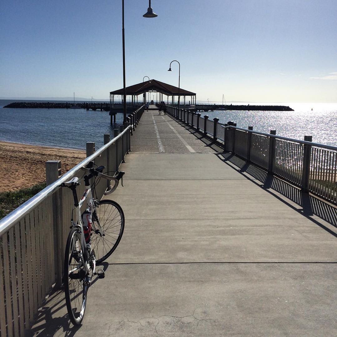 Bicycle leaning against a railing on a long pier leading to a covered shelter over calm water in bright sunlight.