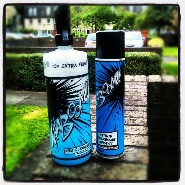 Two blue and black Kaboom cleaning product bottles on a stone wall outdoors, one labeled bike cleaner and one citrus degreaser spray.