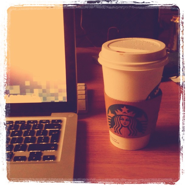 Starbucks paper cup with lid on a wooden desk beside an open laptop.