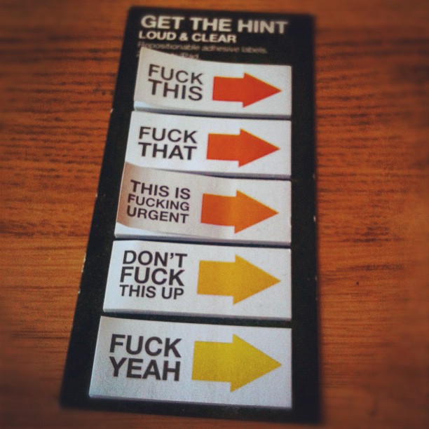 Packet of novelty sticky labels on a wooden table, each with a colored arrow and profanity including “FUCK THIS,” “THIS IS FUCKING URGENT,” and “FUCK YEAH.”
