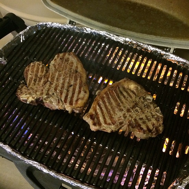 Three steaks grilling on a barbecue with foil-lined edges and flames visible below the grate.
