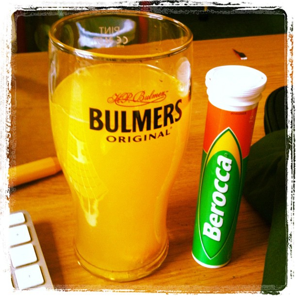 A pint glass of Bulmers cider beside a tube of Berocca on a wooden desk.