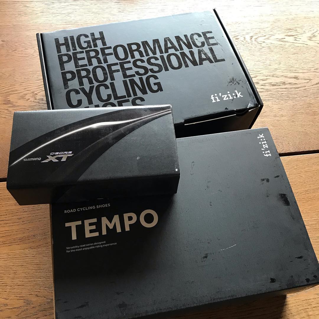 Three black cycling product boxes on a wooden table, including Shimano Deore XT, Fizik, and Tempo road cycling shoes packaging.