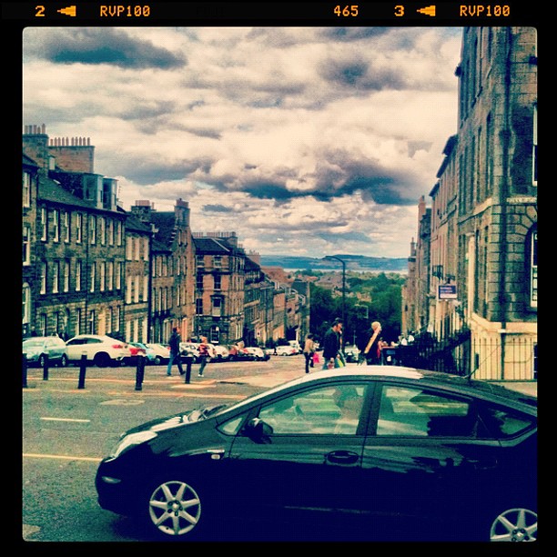Black car parked on a sloping city street lined with stone buildings under a cloudy sky.