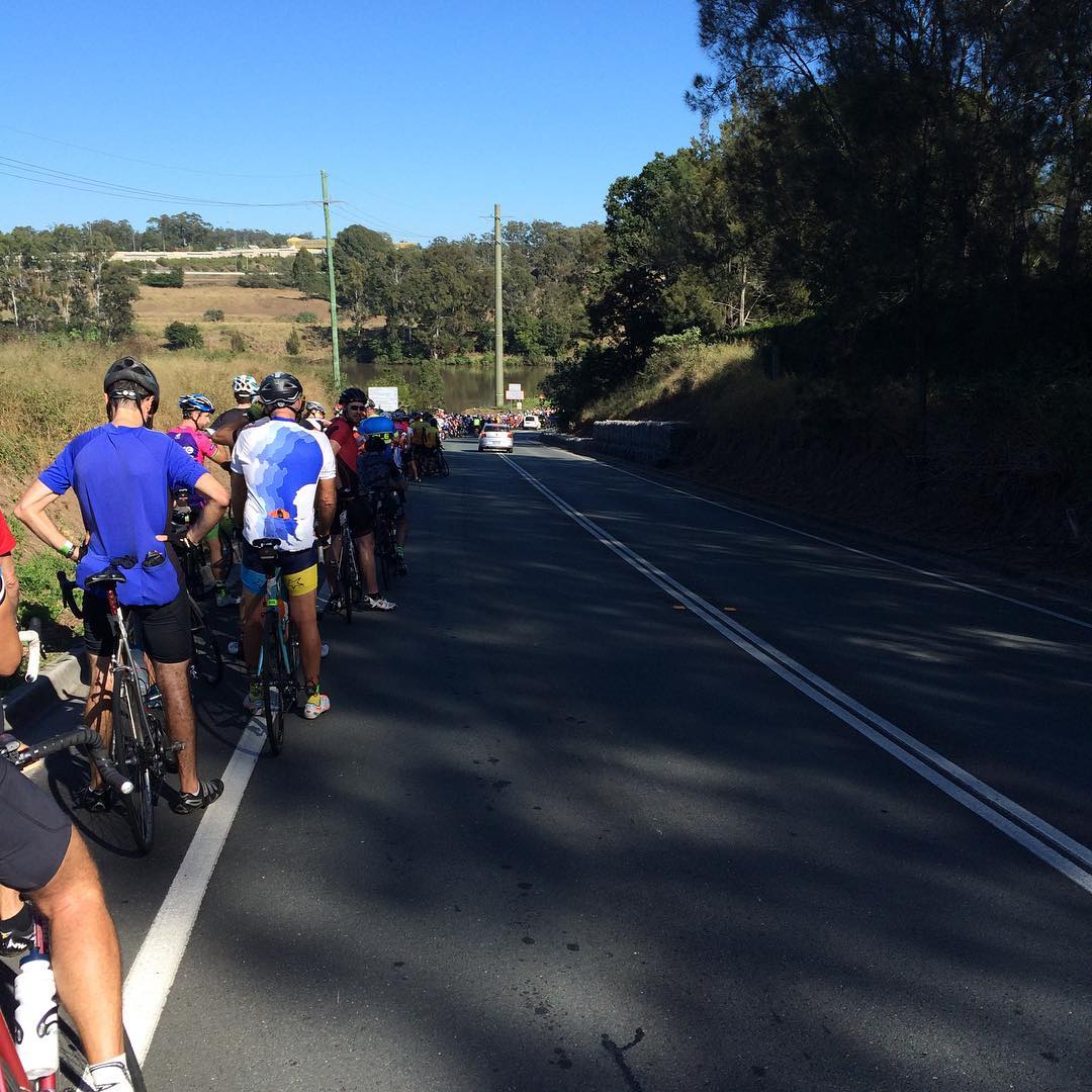 Cyclists in helmets stand with their bikes along the shoulder of a two-lane road bordered by trees under a clear blue sky.