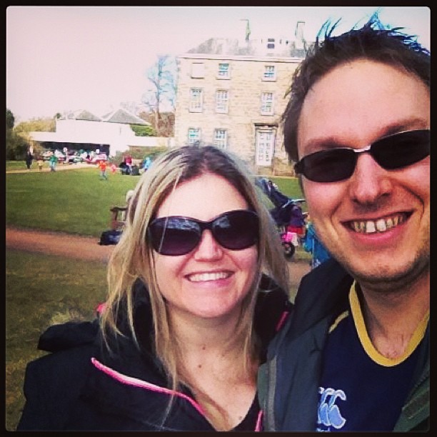 Two smiling people wearing sunglasses pose outdoors in front of a large stone building and grassy lawn.