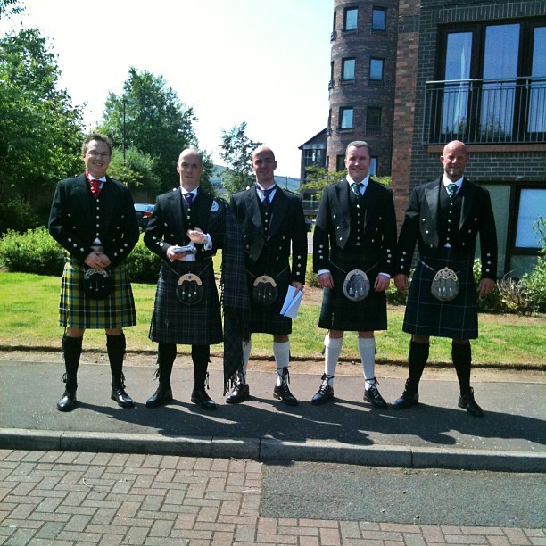 Five men in formal jackets and kilts stand on a sidewalk in sunlight beside a brick apartment building.