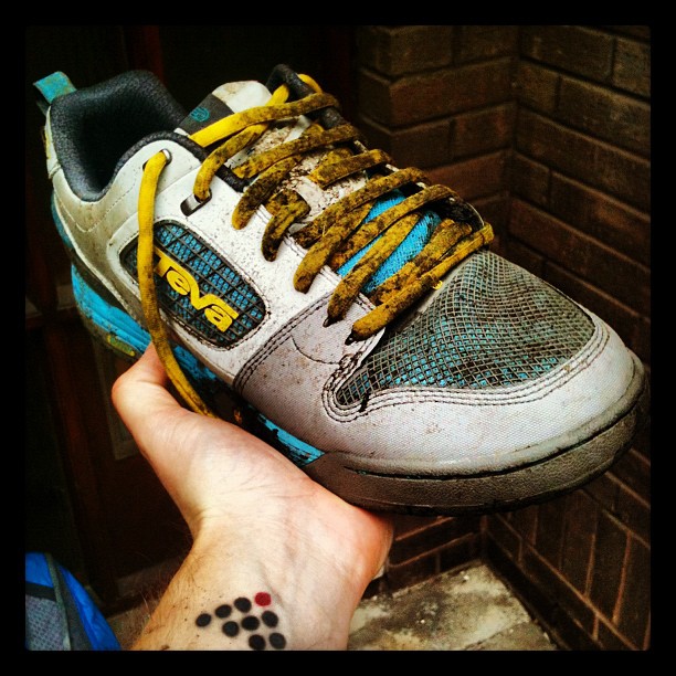 Hand holding a muddy gray, blue, and yellow Teva sneaker with dirty yellow laces against a brick wall.