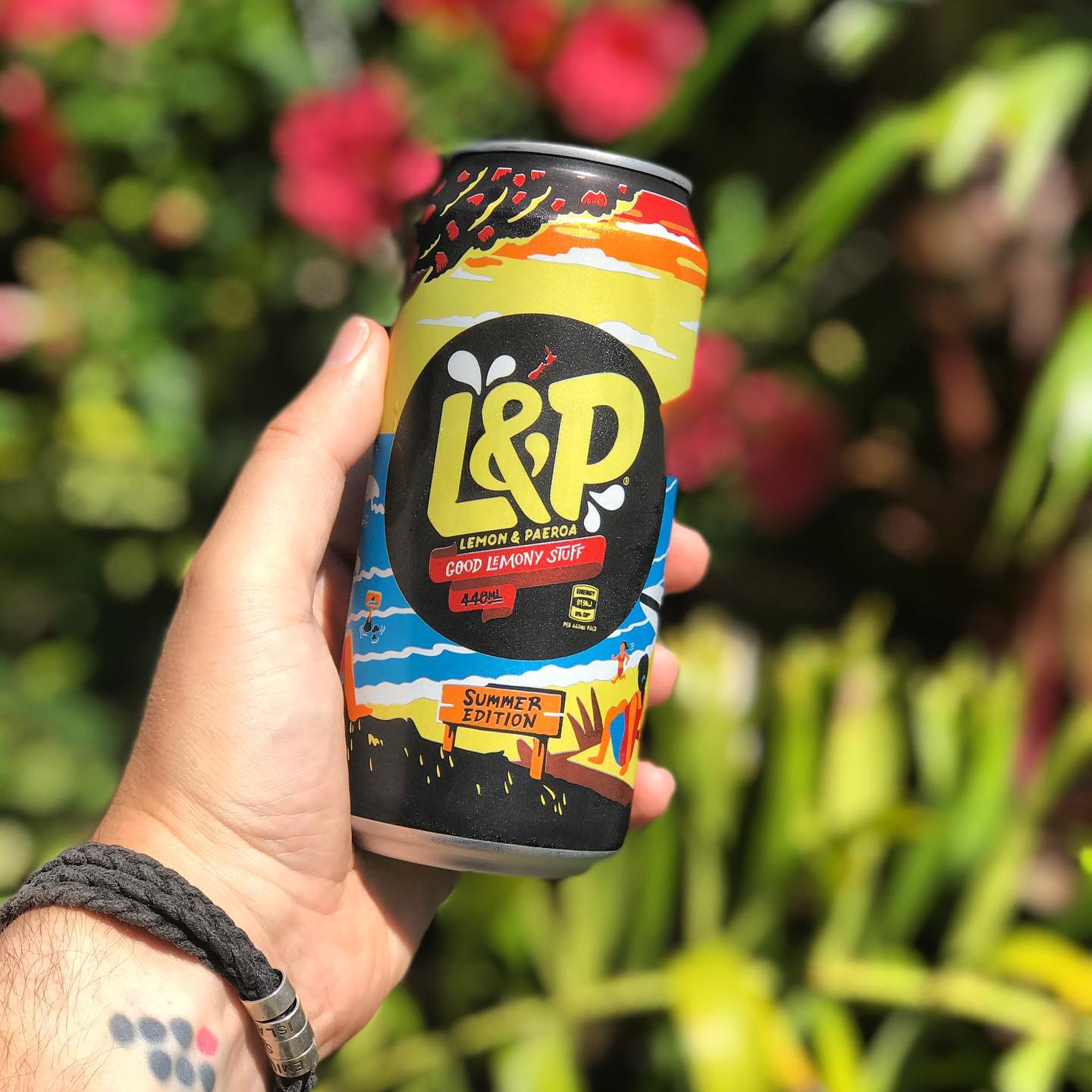 Hand holding a colorful L&P Lemon & Paeroa Summer Edition can outdoors against a blurred background of green leaves and pink flowers.