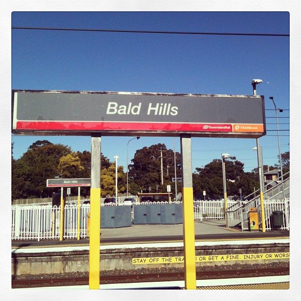 Bald Hills train station sign on a platform with tracks, fencing, trees, and a clear blue sky.