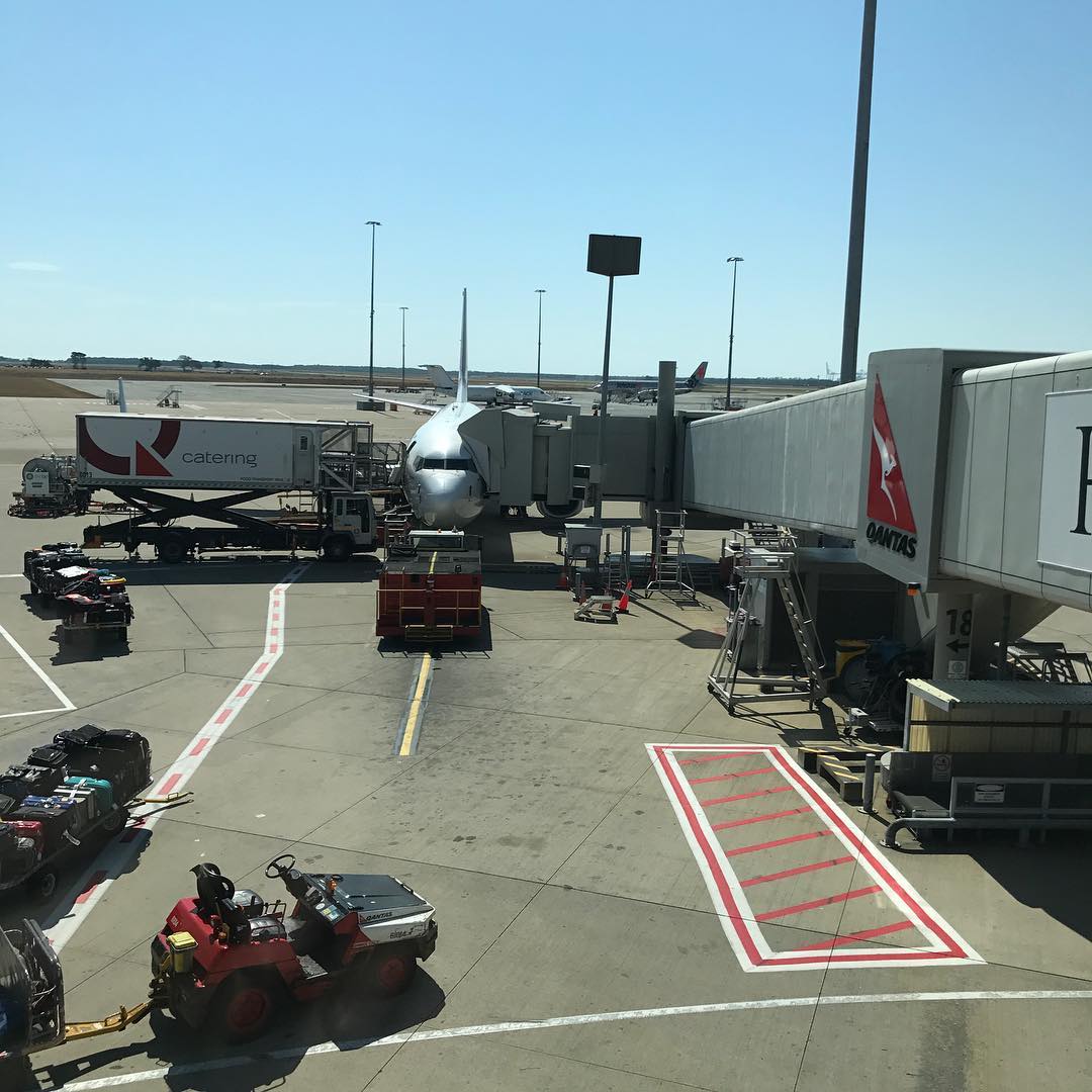 Airplane parked at an airport gate with a jet bridge attached, baggage carts, and a catering truck on the tarmac.
