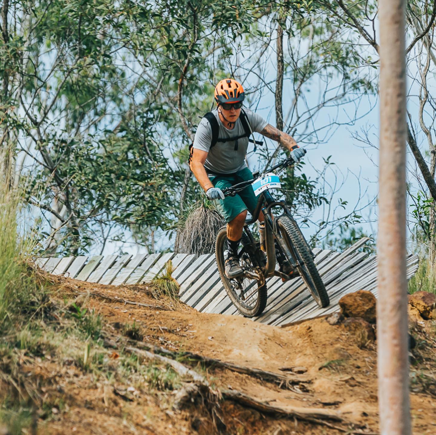 Mountain biker in an orange helmet rides down a wooden banked trail section through a wooded area.