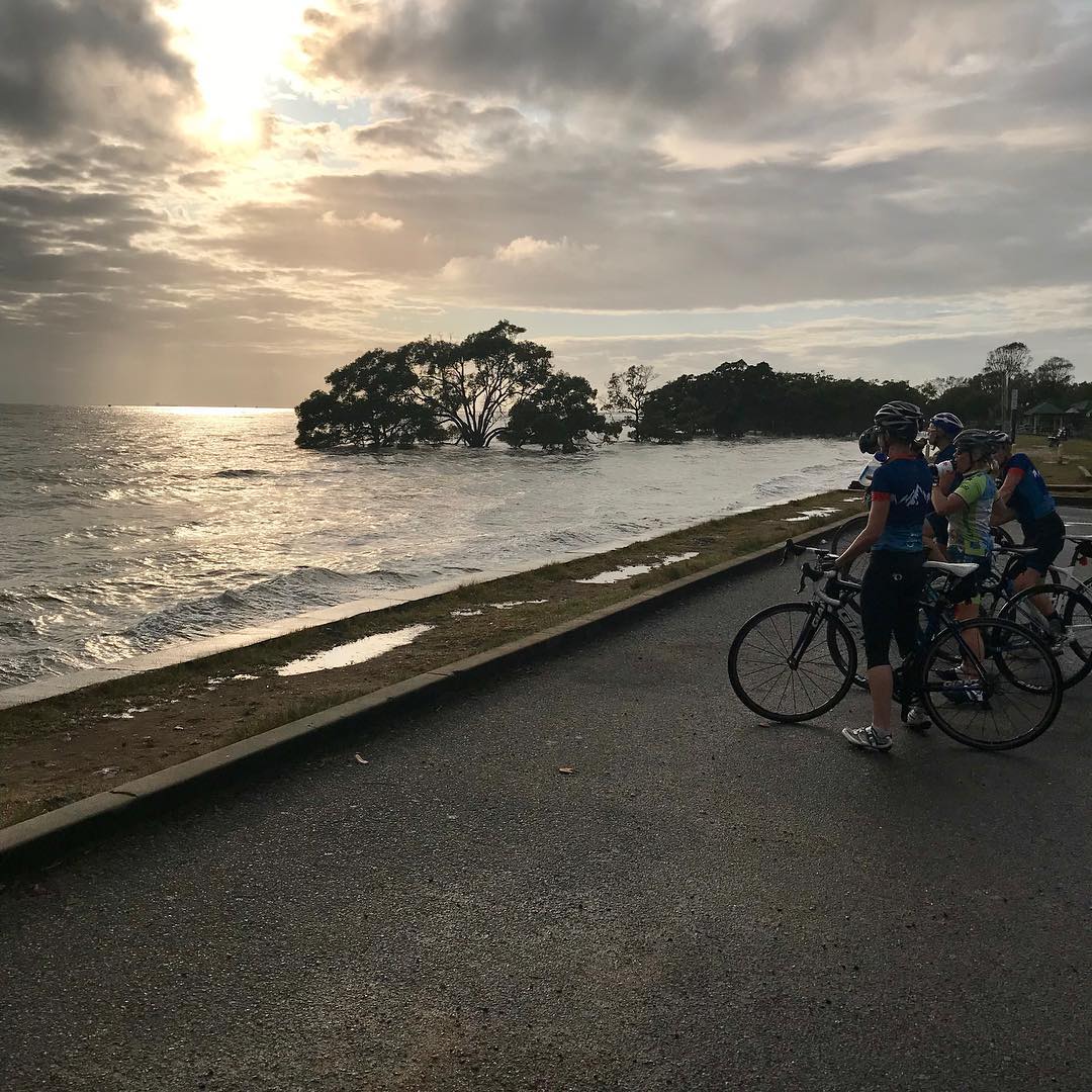 Cyclists with road bikes stand on a wet waterfront path, watching waves and partially flooded trees under a dramatic cloudy sky.