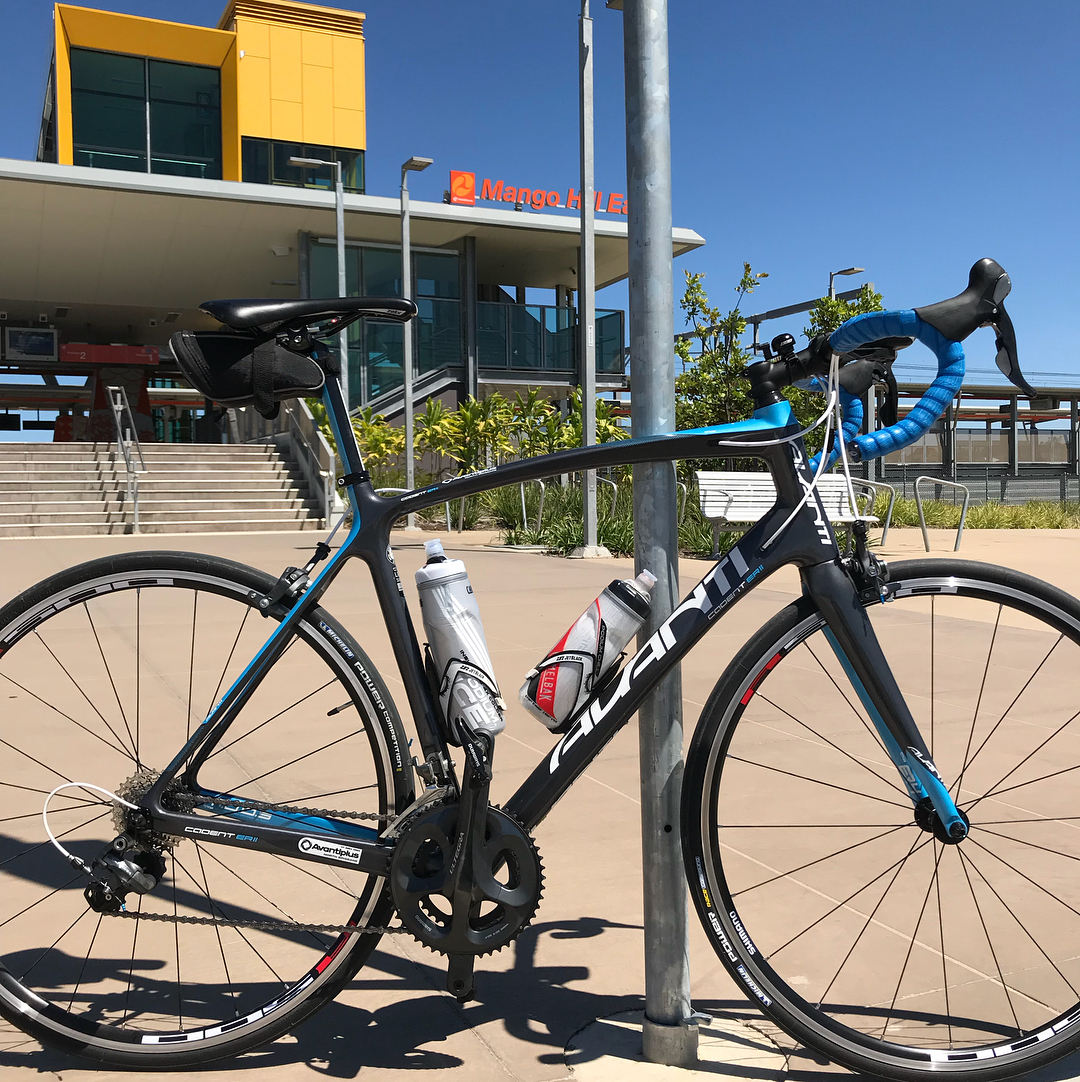 Black and blue road bike locked to a pole outside a modern station building under a clear blue sky.