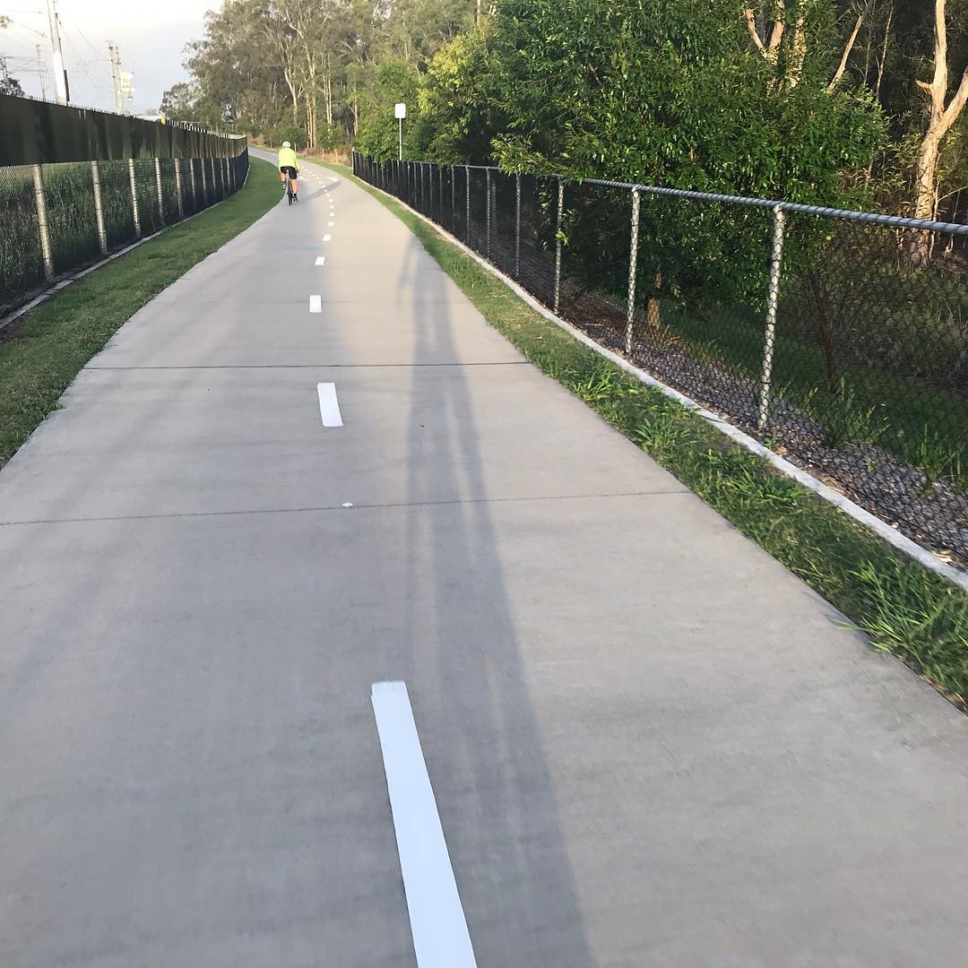Paved shared path with center markings, fenced on both sides, with a cyclist ahead and long shadows cast across the path.
