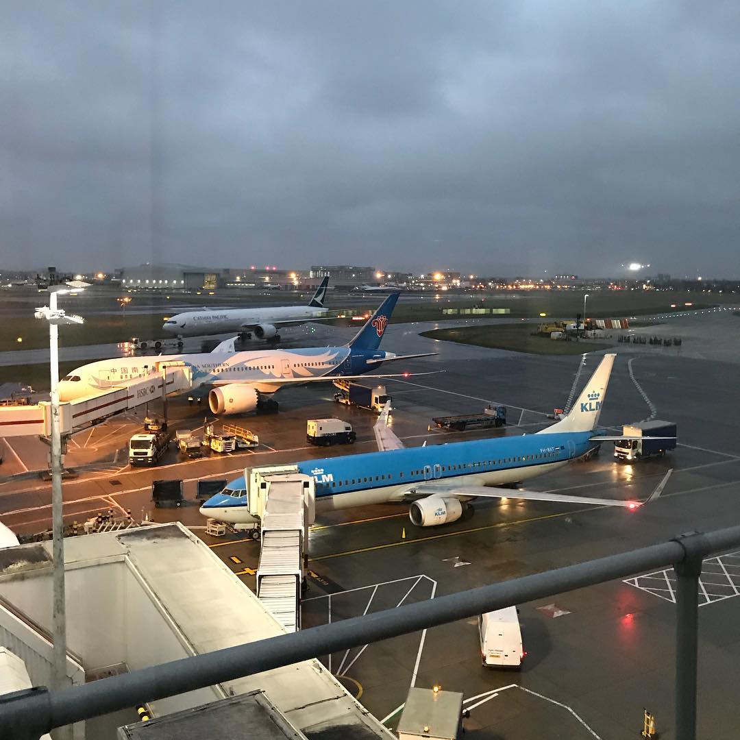 Three airplanes parked at airport gates on a wet tarmac at dusk, with service vehicles and runway lights under a cloudy sky.