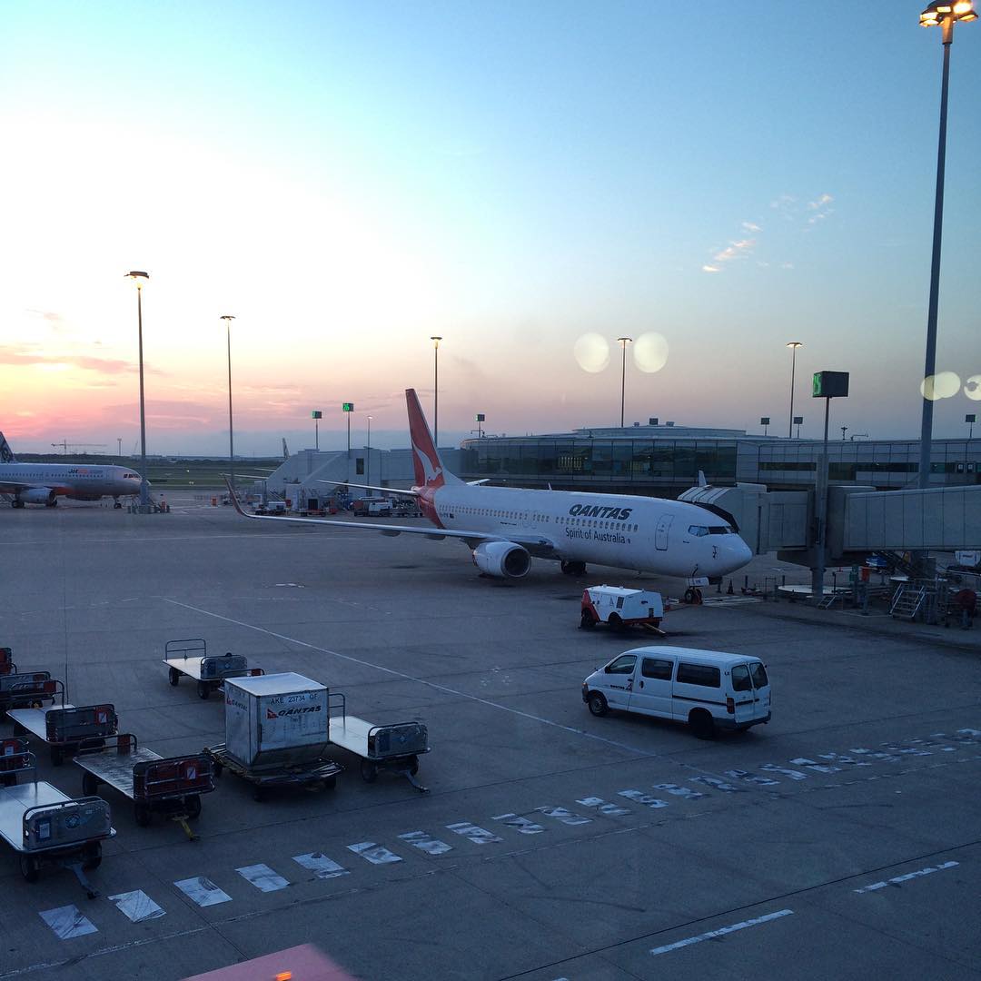 Qantas plane parked at an airport gate at dusk, with baggage carts, service vehicles, terminal buildings, and tall light poles nearby.