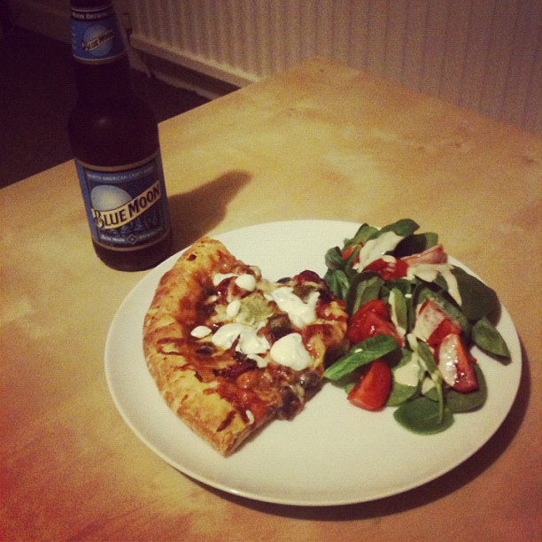 Plate with two slices of pizza and a salad beside a bottle of Blue Moon beer on a wooden table.