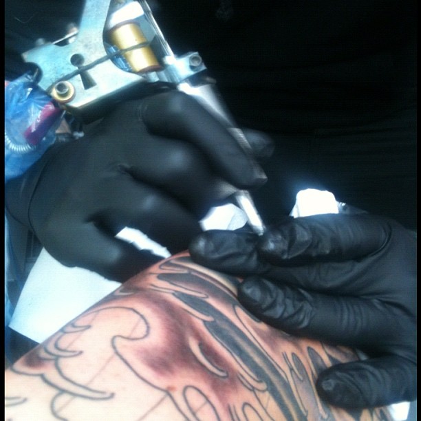 Black-gloved hands use a tattoo machine to ink and shade a colorful tattoo on a person's arm.