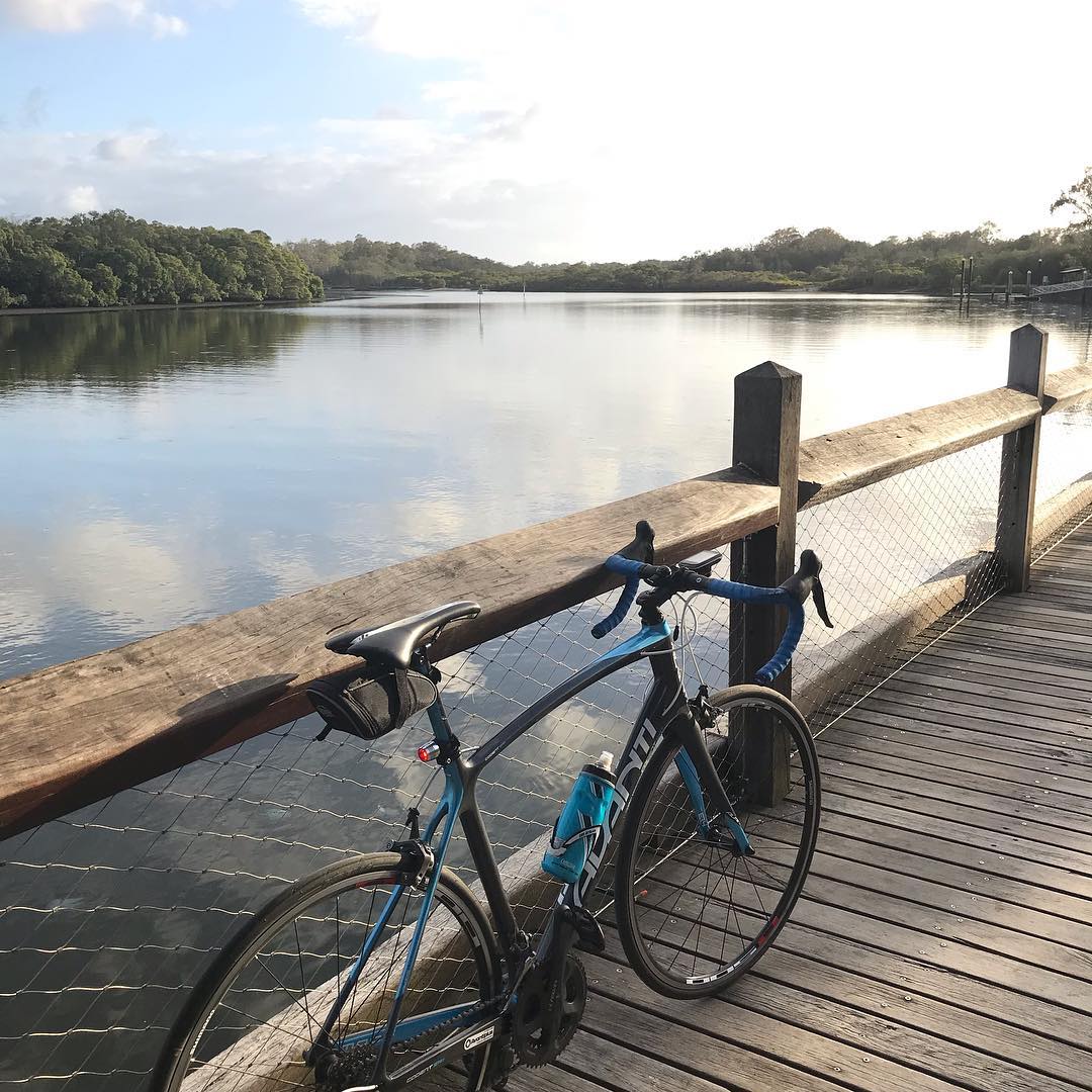 Road bike parked against a wooden boardwalk railing beside a calm river, with trees and bright sky reflected on the water.