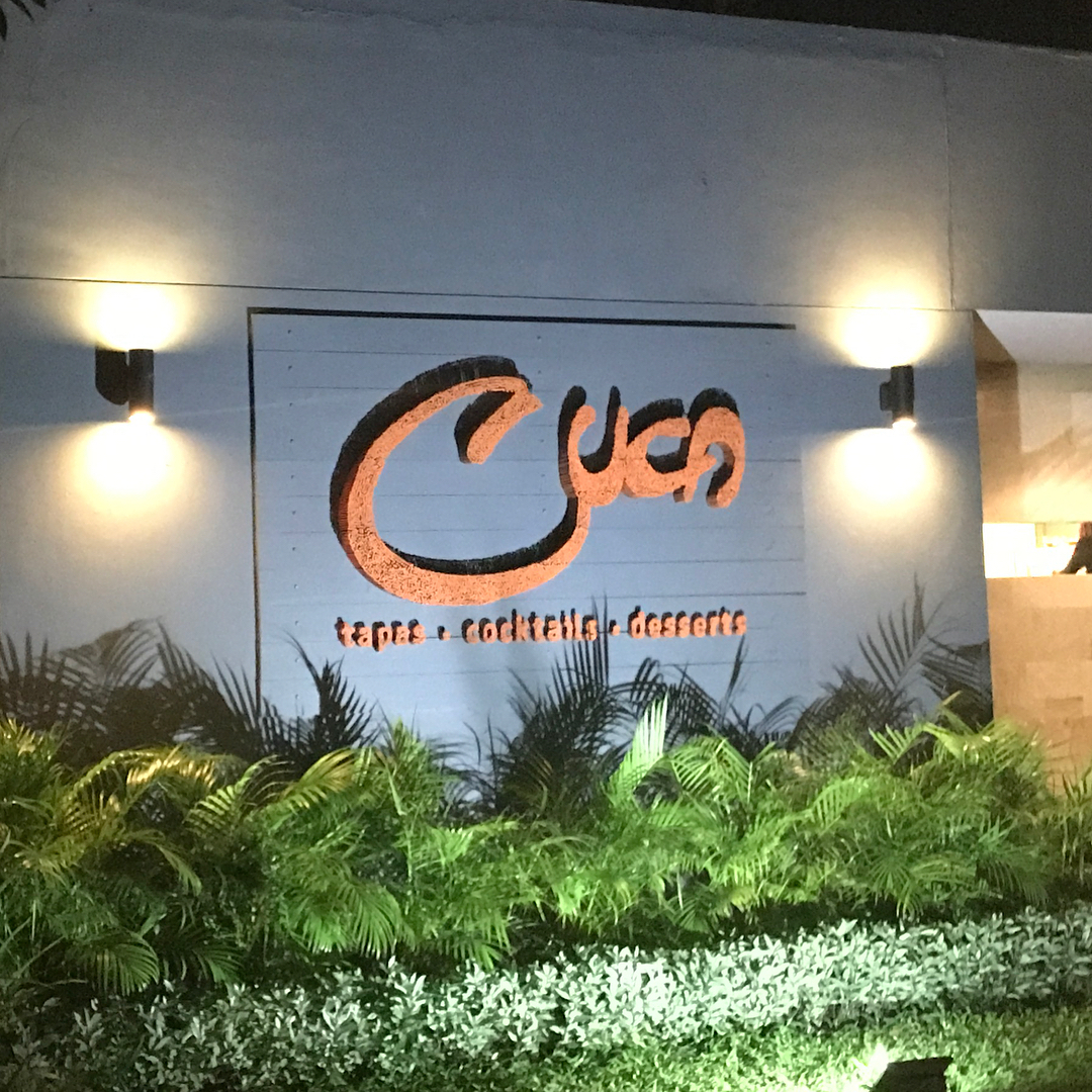 Lit restaurant sign reading "Cya" with "tapas, cocktails, desserts" on a white wall above tropical plants at night.
