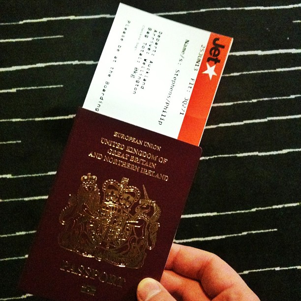 Hand holding a UK passport with a Jetstar boarding pass partially visible against a black striped background.