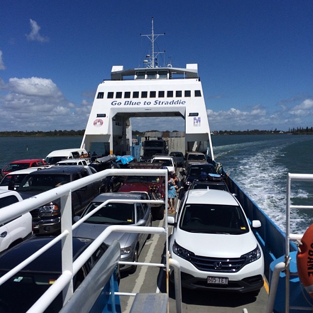 Car ferry deck packed with parked vehicles under a blue sky, with “Go Blue to Straddie” on the front and white wake trailing behind.