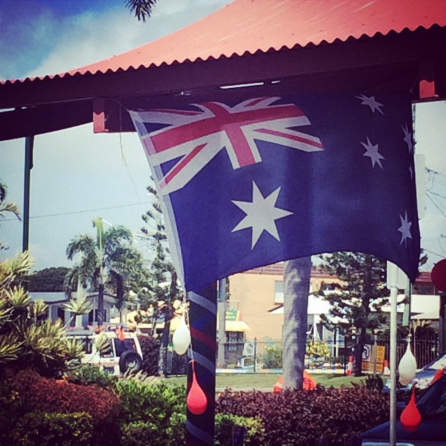 Australian flag hanging beneath a red corrugated roof with trees, shrubs, and street buildings in the background.