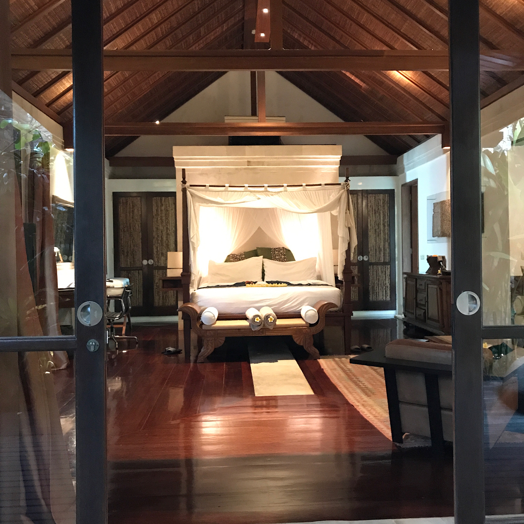 Warmly lit bedroom with a canopy bed, polished wood floors, high vaulted ceiling, and dark wood furniture seen through glass doors.