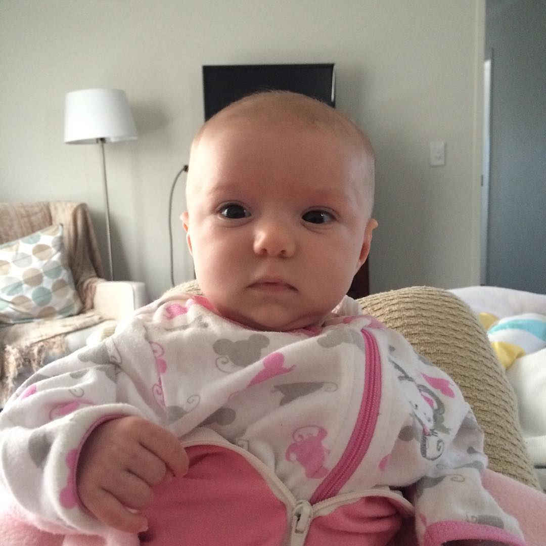 Baby in a white and pink patterned outfit sitting indoors, looking directly at the camera.
