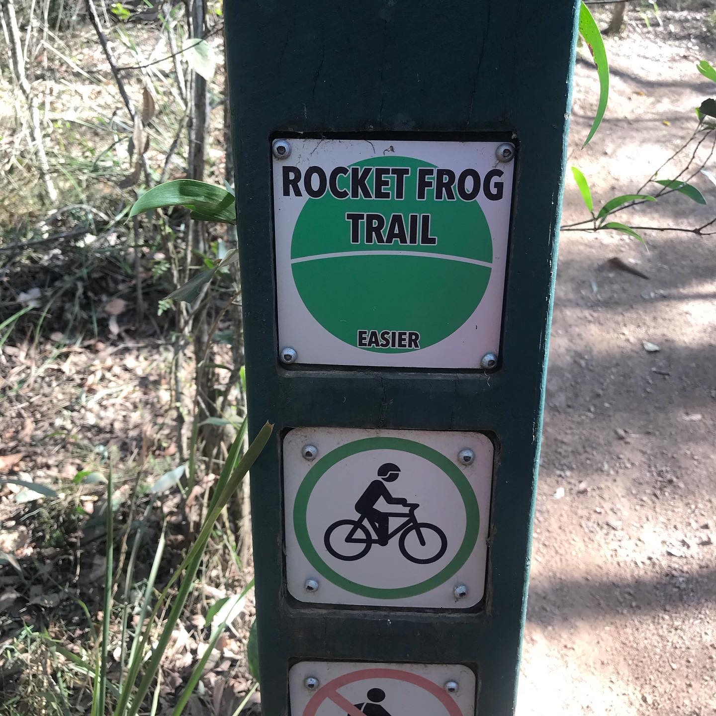 Signpost on a wooded trail reading “Rocket Frog Trail” with a green easier rating and a bicycle symbol below.