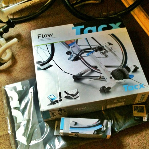 Boxed Tacx Flow bike trainer and accessories on a carpet, with part of a bicycle wheel visible above.