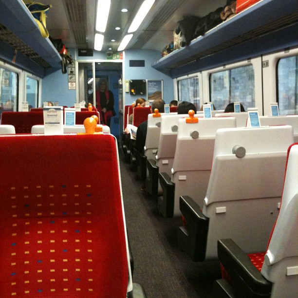 Interior of a train car with red and white seats, overhead luggage racks, and a few passengers seated down the aisle.
