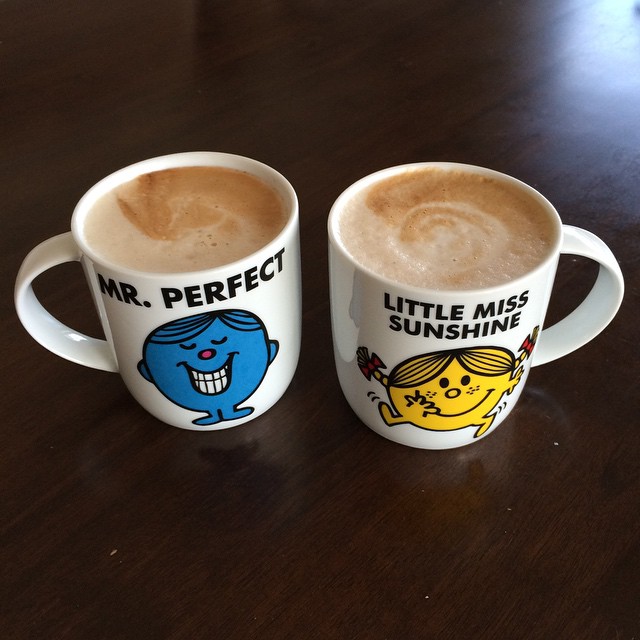 Two mugs of coffee on a dark wooden table, labeled “Mr. Perfect” and “Little Miss Sunshine” with cartoon characters.