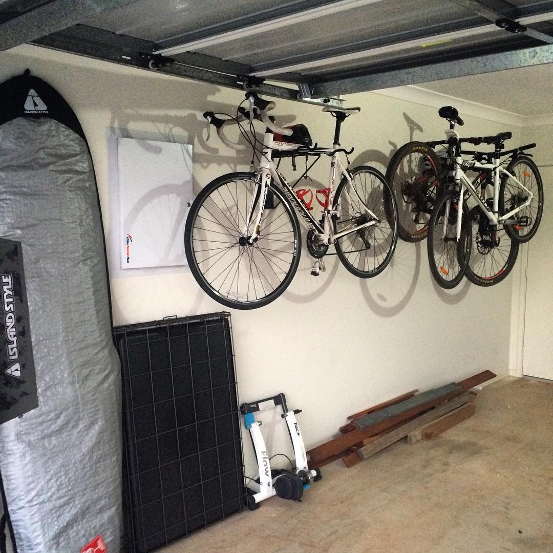 Three bicycles hang from wall mounts in a garage, with a folded table, exercise stand, and stacked wood below.