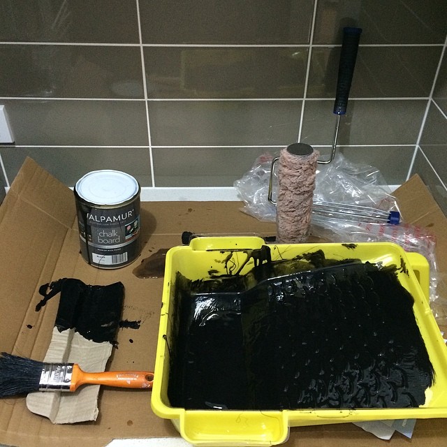Paint tray filled with black paint, a brush, paint roller, and a can of chalkboard paint on cardboard against a tiled wall.