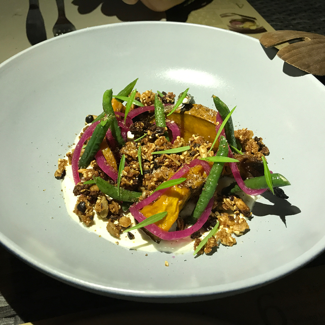 Roasted squash with green beans, pickled red onion, herbs, and crunchy granola-like topping over a white sauce in a wide bowl.