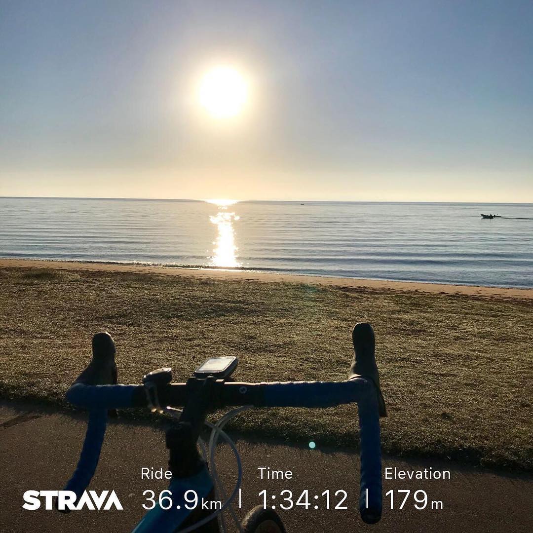 Bicycle handlebars in the foreground facing a calm beach and bright sun over the water, with Strava ride stats overlaid at the bottom.